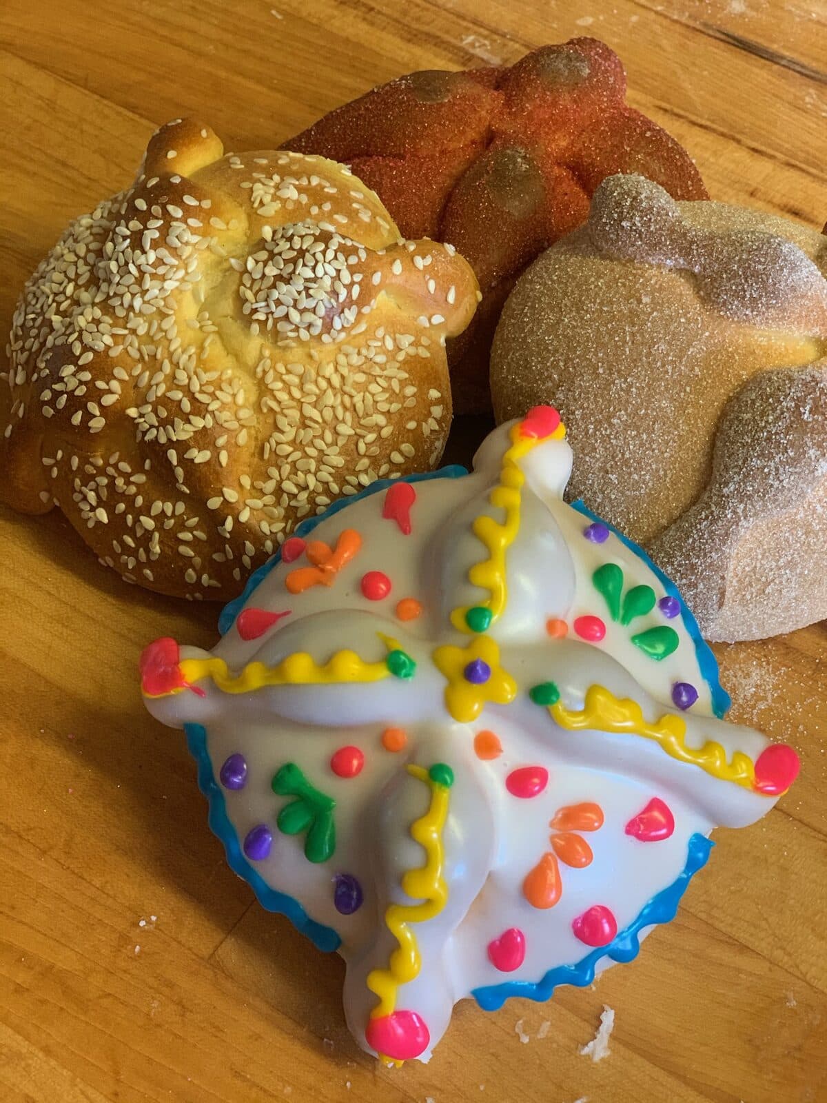 Cuatro estilos de pan de muerto: con ajonjolí, con azúcar, sencillo y con glaseado de colores, sobre una mesa de panadería